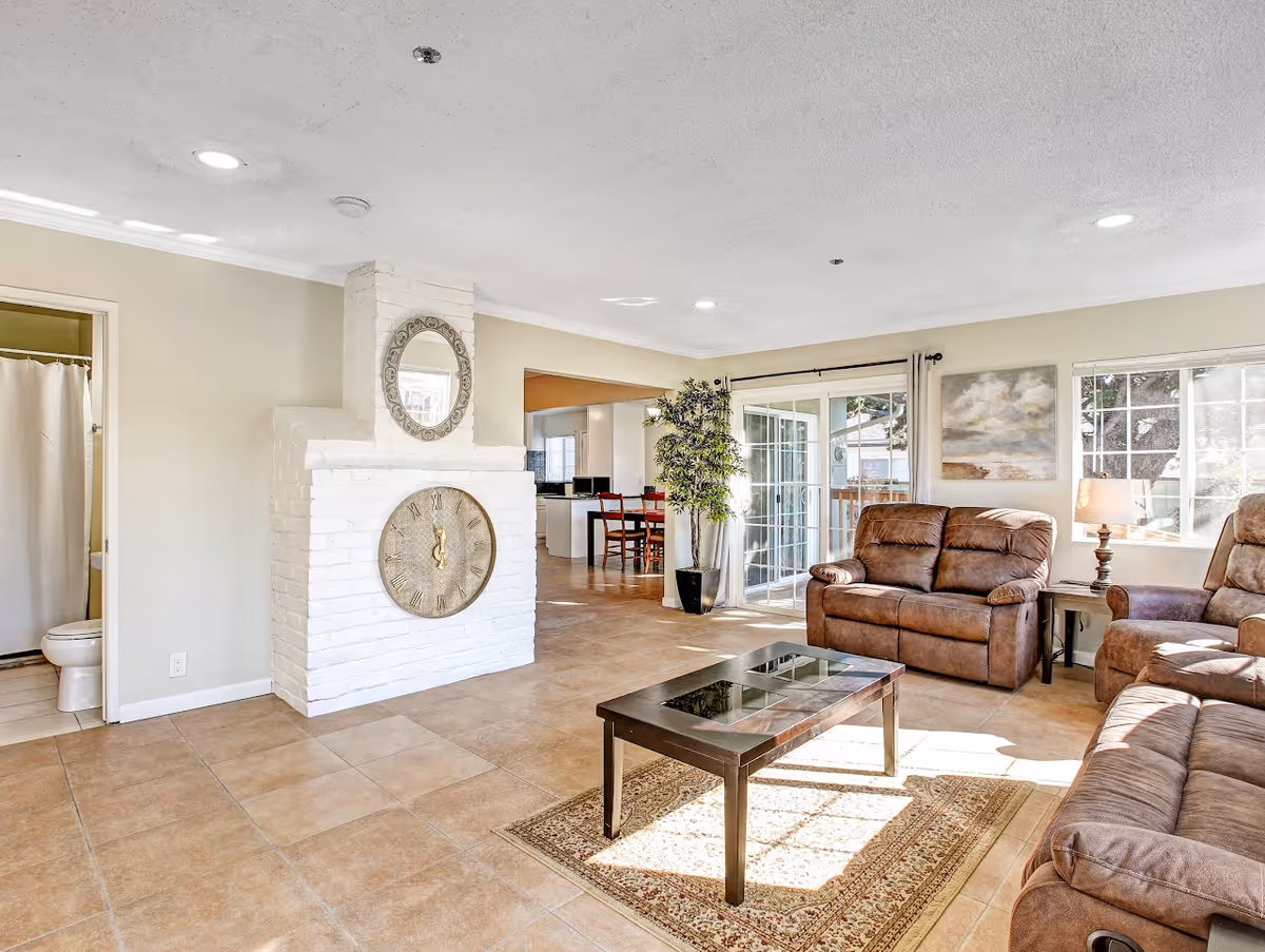 Bright living room with brown leather recliners, a glass-top coffee table on a rug, a white brick fireplace with decorative clock and mirror, and sliding glass doors.