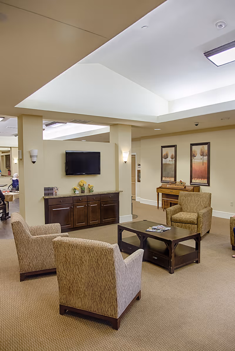 A cozy assisted living facility common area with beige walls and carpet. The room features three upholstered armchairs arranged around a dark wooden coffee table with magazines on it. A flat-screen TV is mounted on the wall above a cabinet with decorative flowers. Two framed landscape paintings hang on the wall above a small wooden piano. The ceiling has recessed lighting and a skylight.