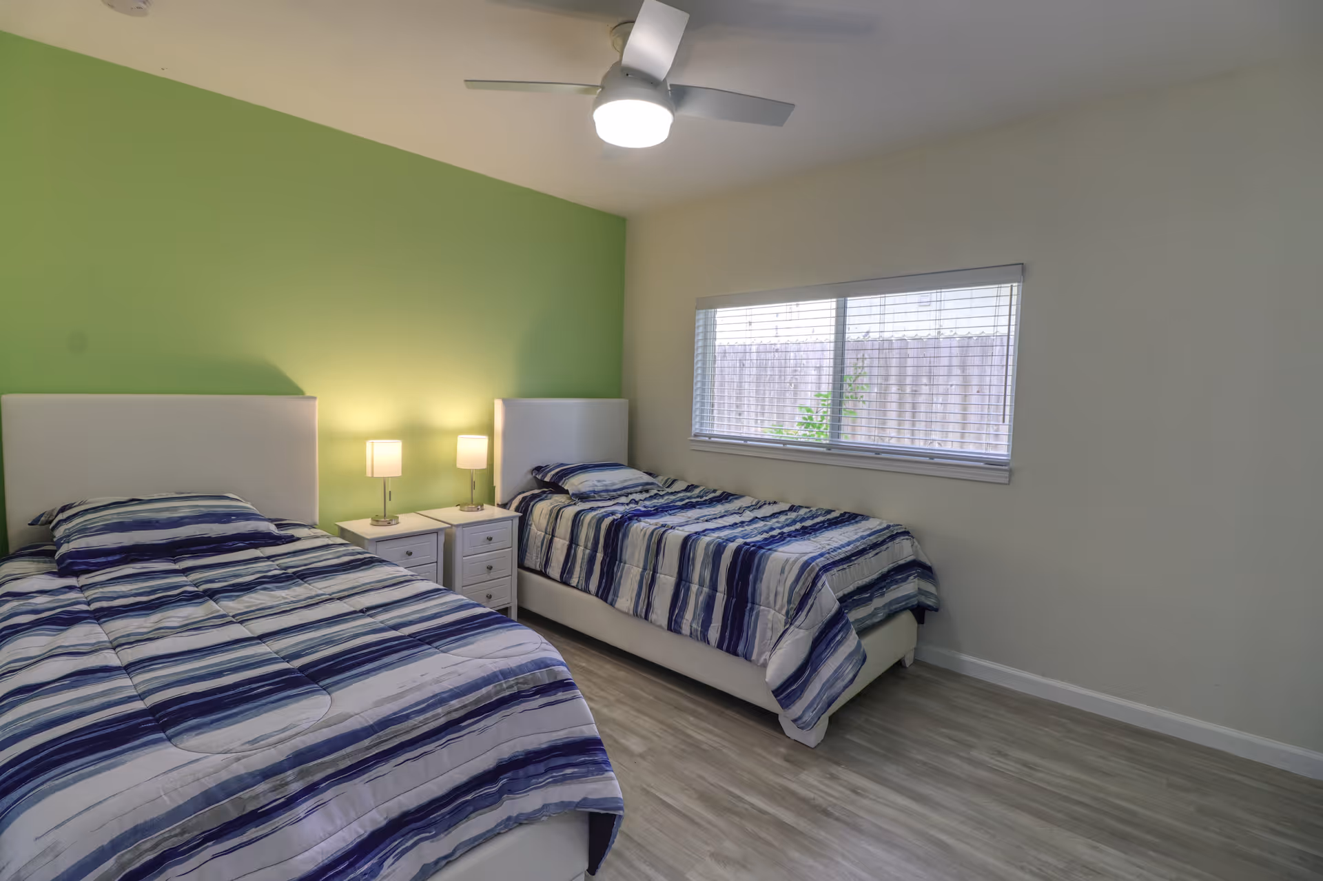 A bedroom with two single beds featuring white headboards and blue striped bedding. Between the beds is a white nightstand with two lamps. The room has a green accent wall behind the beds, a ceiling fan with a light, a window with blinds, and light-colored wood flooring.