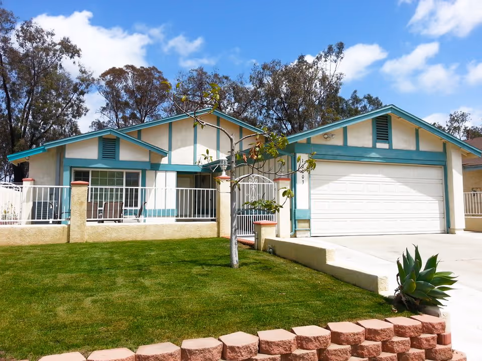 Single-story beige house with teal trim, attached two-car garage, front lawn and white fence under a blue sky.