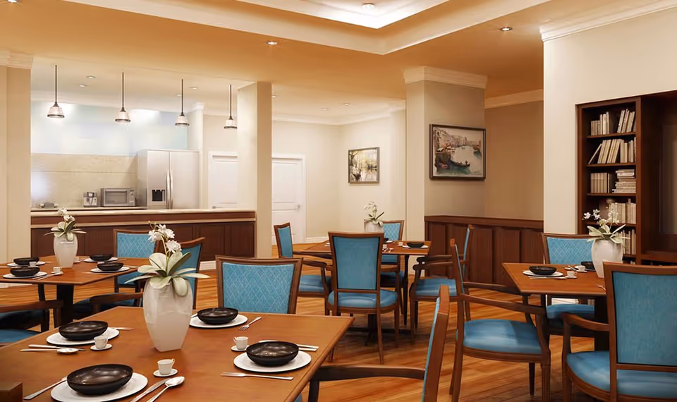A dining room with several wooden tables set with black bowls, white plates, small cups, and silverware. Each table has a white vase with white flowers. The chairs have blue cushioned seats and backs. In the background, there is a kitchen area with a refrigerator, microwave, and coffee maker. The walls are decorated with framed paintings and a built-in bookshelf filled with books.
