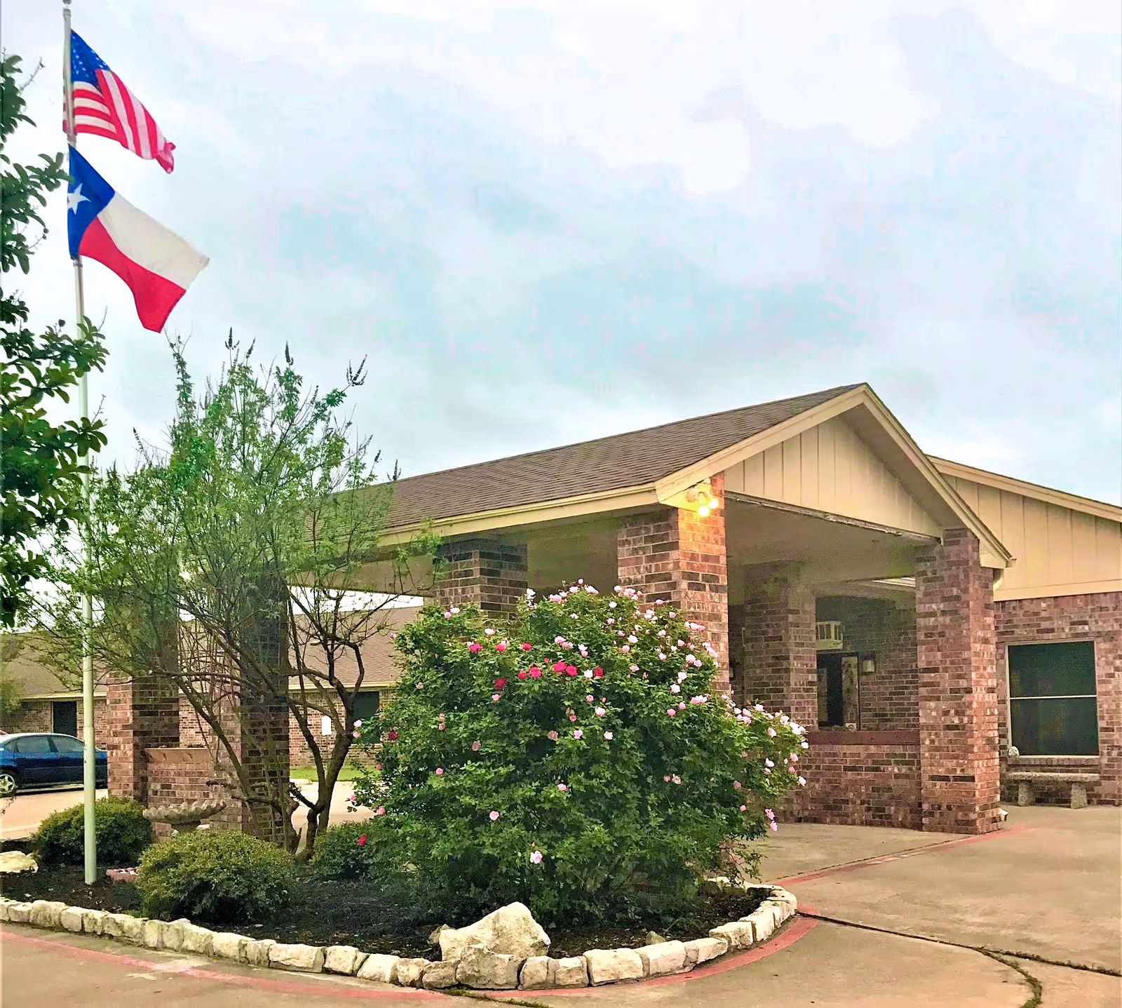 Front entrance of a single-story brick nursing facility with a covered porte-cochere, flags flying, and landscaped shrubs and flowering bushes.