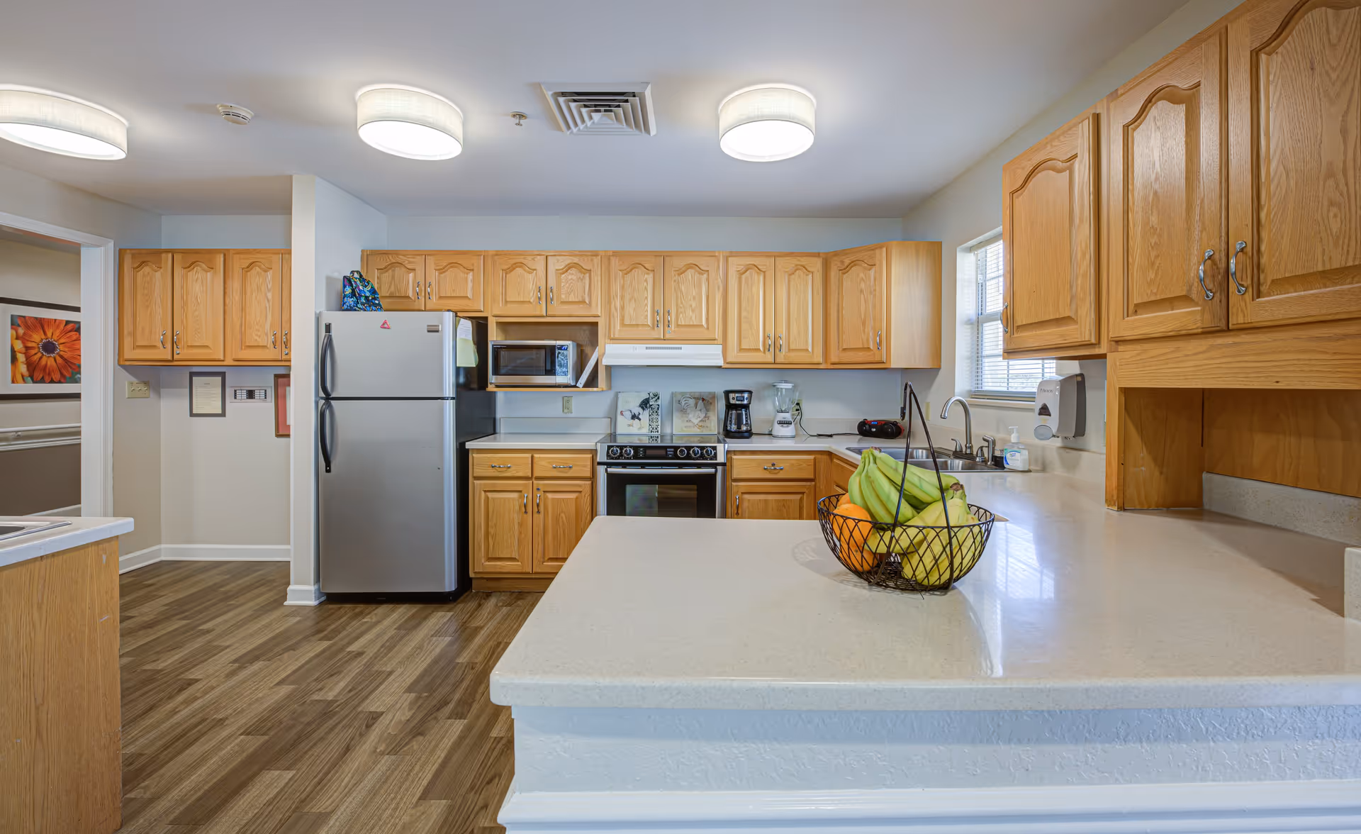 A clean and well-lit kitchen with wooden cabinets, a stainless steel refrigerator, a microwave, an electric stove with an oven, a coffee maker, a blender, and a sink under a window. A basket of fruit is placed on the light-colored countertop island in the foreground. The floor has a wood-like finish, and there are ceiling lights providing illumination.