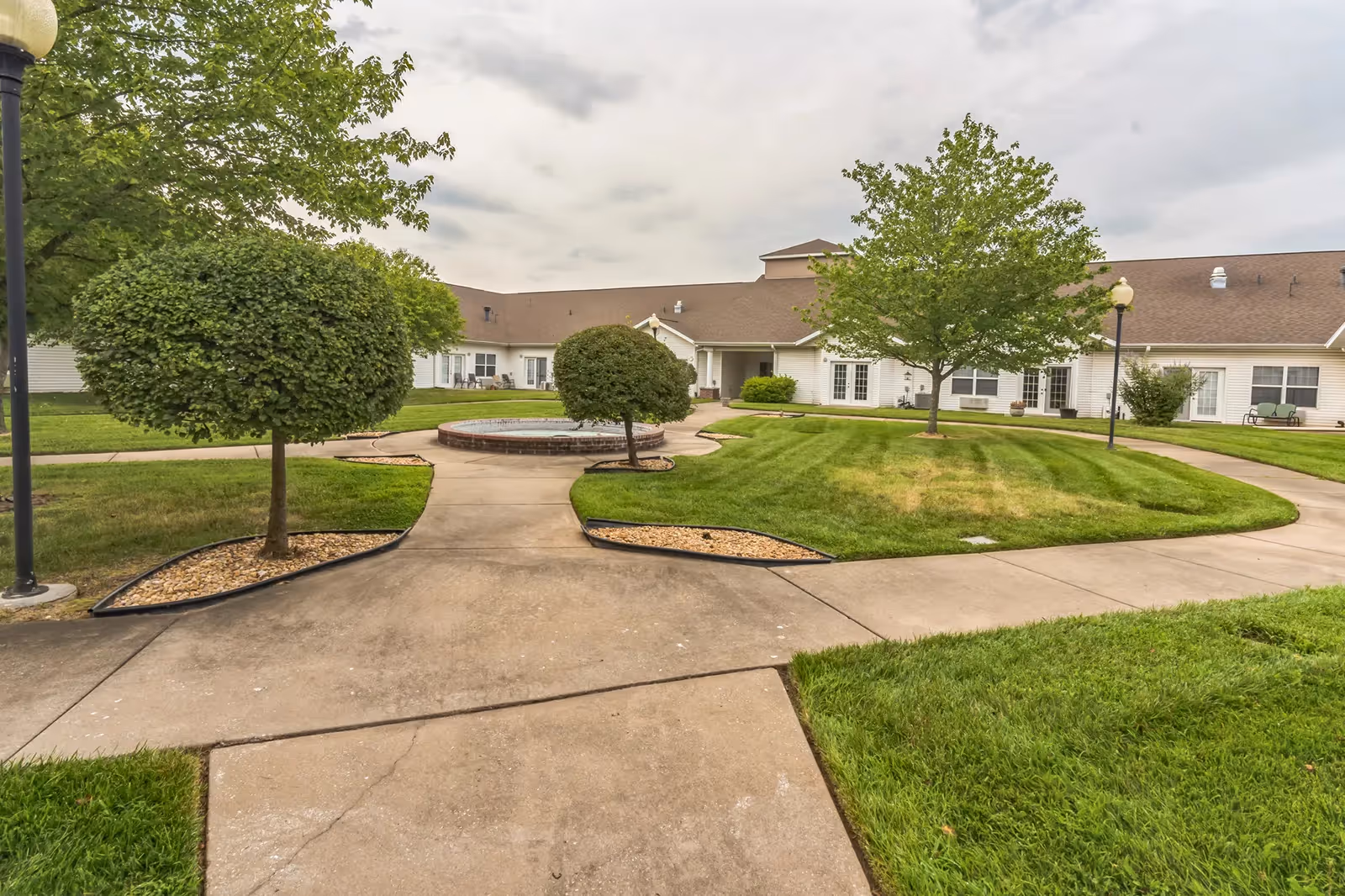 Outdoor courtyard area with well-maintained green grass, trimmed bushes, and trees. Concrete walkways lead to a circular water fountain in the center. The background shows a single-story building with white siding and multiple windows and doors. The sky is cloudy.
