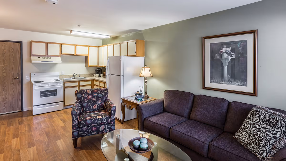 A cozy living room and kitchen area in a senior living facility. The living room features a dark purple sofa with a patterned pillow, a floral armchair, a glass coffee table with decorative balls, and a wooden side table with a lamp and small decor items. The kitchen has wooden cabinets, a white refrigerator, a white stove with an oven, a double sink, and a coffee maker. The walls are painted light green and beige, and there is a framed black and white picture hanging above the sofa.