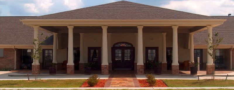Front entrance of a single-story brick building with a covered portico supported by columns, double doors, and a walkway with small landscaped beds.