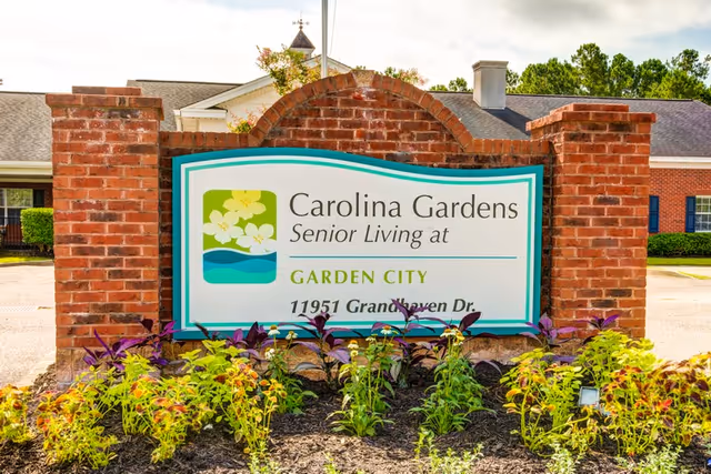 A brick signpost with a white and green sign that reads 'Carolina Gardens Senior Living at Garden City 11951 Grandhaven Dr.' surrounded by plants and flowers, with a building and trees in the background.