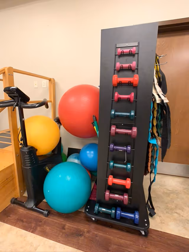 A small exercise area with a stationary bike, several large colorful exercise balls, and a black rack holding multiple pairs of colorful dumbbells. There are also some resistance bands hanging on the side of the rack. The room has a wooden floor section and a beige wall with a wooden door in the background.