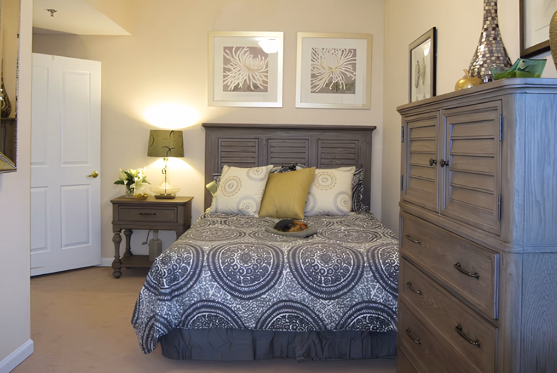 A cozy bedroom with a patterned bedspread, wooden headboard and dresser, a nightstand with a lamp, and framed artwork above the bed.