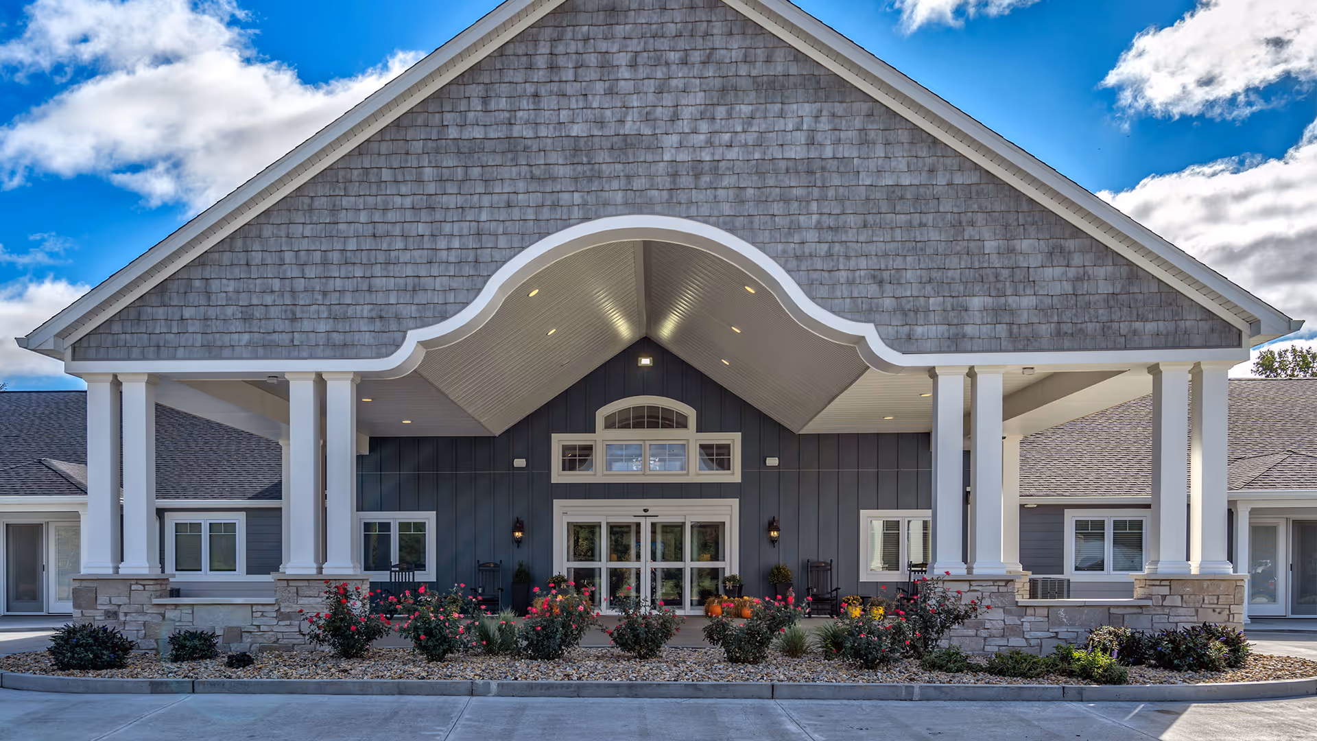 Front exterior view of Villas of Holly Brook Assisted Living & Memory Care facility in Bethalto, IL, featuring a large covered entrance with white columns, stone accents, and landscaped flower beds under a partly cloudy blue sky.