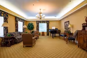 Spacious senior living room with upholstered sofas and chairs, a grand piano, chandelier, patterned carpet, and windows with floral curtains.