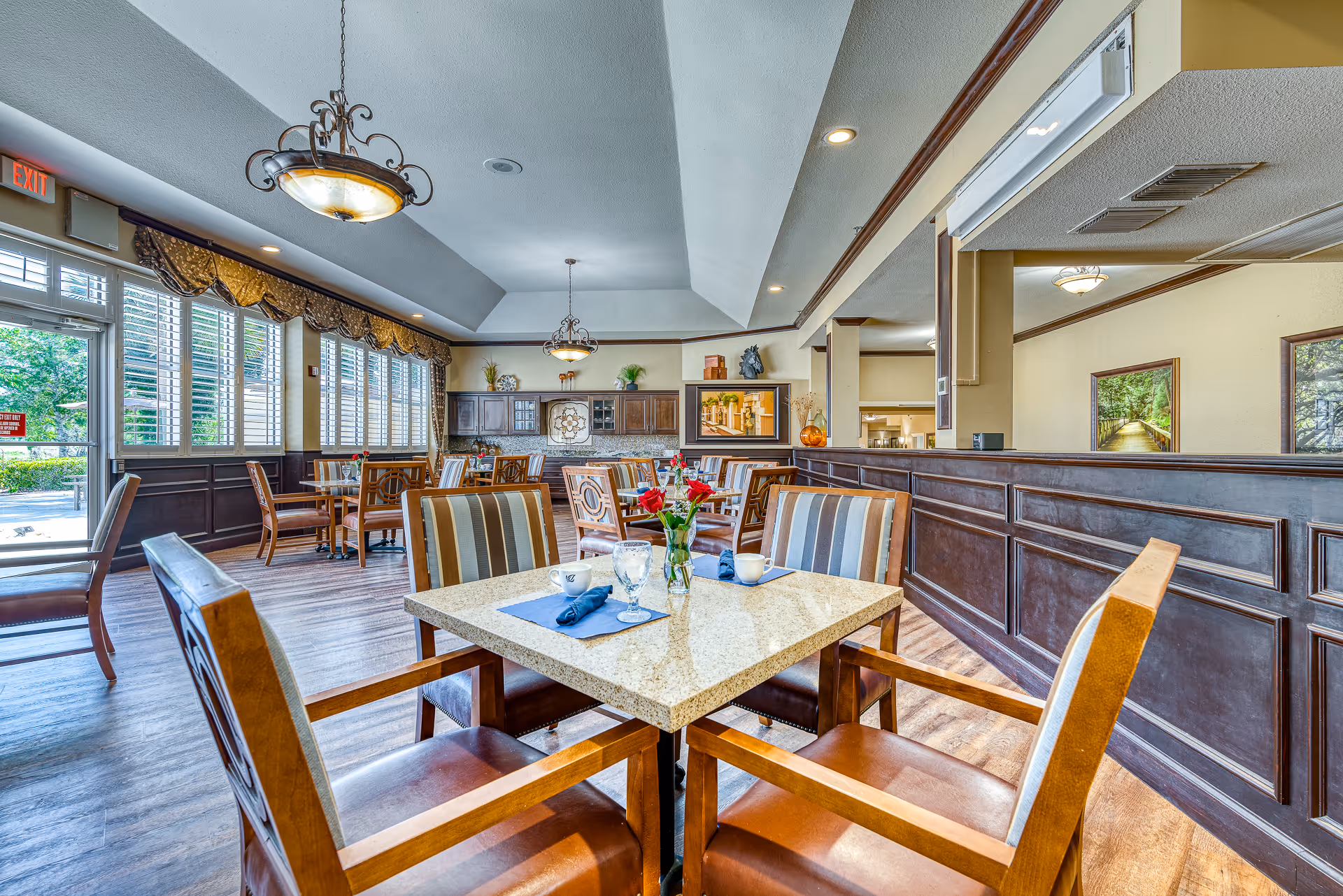 Dining room with a set table in the foreground, wooden chairs, large windows with shutters, and decorative chandeliers.
