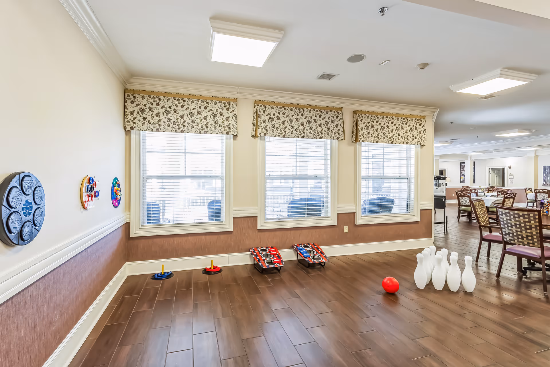 A recreational area inside a senior living facility with wooden flooring and three large windows with floral valances. The room features indoor games including a set of bowling pins with a red ball, two cornhole boards, and ring toss games mounted on the wall. In the background, there are dining tables and chairs arranged in a dining area.