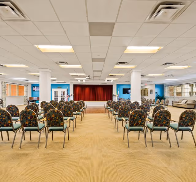 A large room set up with rows of floral-patterned chairs facing a small stage with a red curtain. The room has a beige carpet, white ceiling with fluorescent lights, and white pillars. There are windows along the right side and a seating area with sofas in the back right corner.