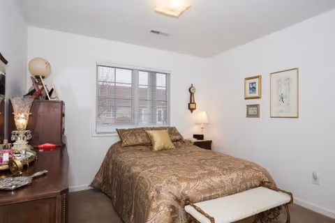 A bedroom with a double bed covered in a brown patterned bedspread and a gold pillow. There is a small bench at the foot of the bed. On the left side, there is a wooden dresser with decorative items including a globe and a lamp. A window with blinds is centered on the back wall. On the right wall, there are three framed pictures and a wall clock above a small nightstand with a lamp.