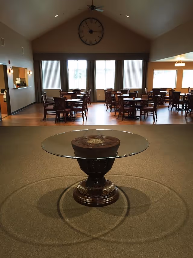 Interior view of a dining room in a senior living facility with multiple round wooden tables and chairs arranged on a wooden floor. In the foreground, there is a decorative round glass table with a carved wooden base on a carpeted floor. Large windows with vertical blinds allow natural light into the room, and a large wall clock is mounted on the beige wall above the windows.