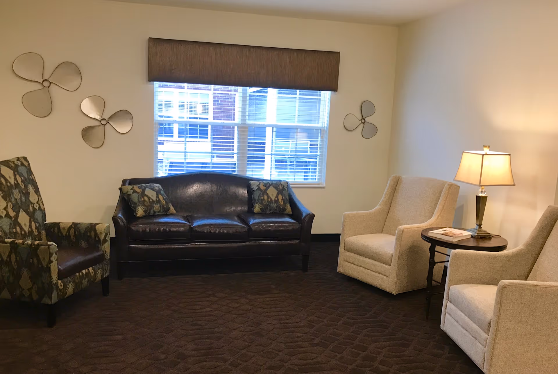 A cozy living room with a dark leather sofa, two beige armchairs, and one patterned armchair arranged around a small round wooden side table with a lamp and a magazine. The room has a window with blinds and a brown valance, and three decorative metal wall hangings shaped like propellers on the walls.