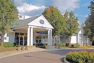 Exterior view of a white building with a green roof and large white columns at the entrance, surrounded by trees and landscaping under a partly cloudy sky.