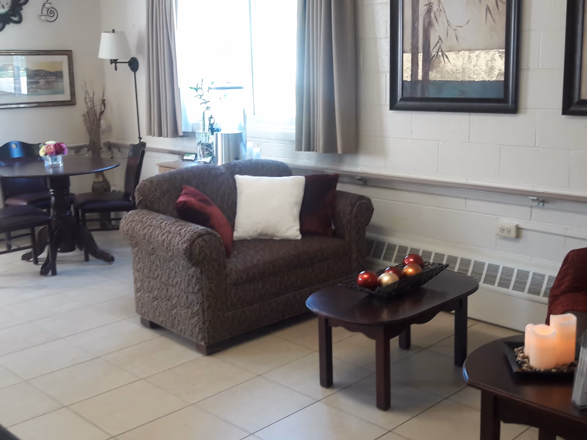 A cozy living room area with a patterned loveseat adorned with two red pillows and one white pillow. In front of the loveseat is a dark wooden coffee table with decorative balls in a black tray. To the right, there is a small side table with three lit candles. In the background, there is a round dining table with four chairs, a wall lamp, framed pictures, and a window with beige curtains letting in natural light.