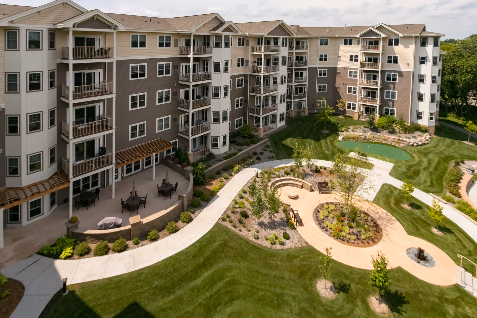 A multi-story senior living facility building with balconies overlooking a landscaped outdoor area featuring walking paths, seating areas, a small putting green, and well-maintained lawns and plants.