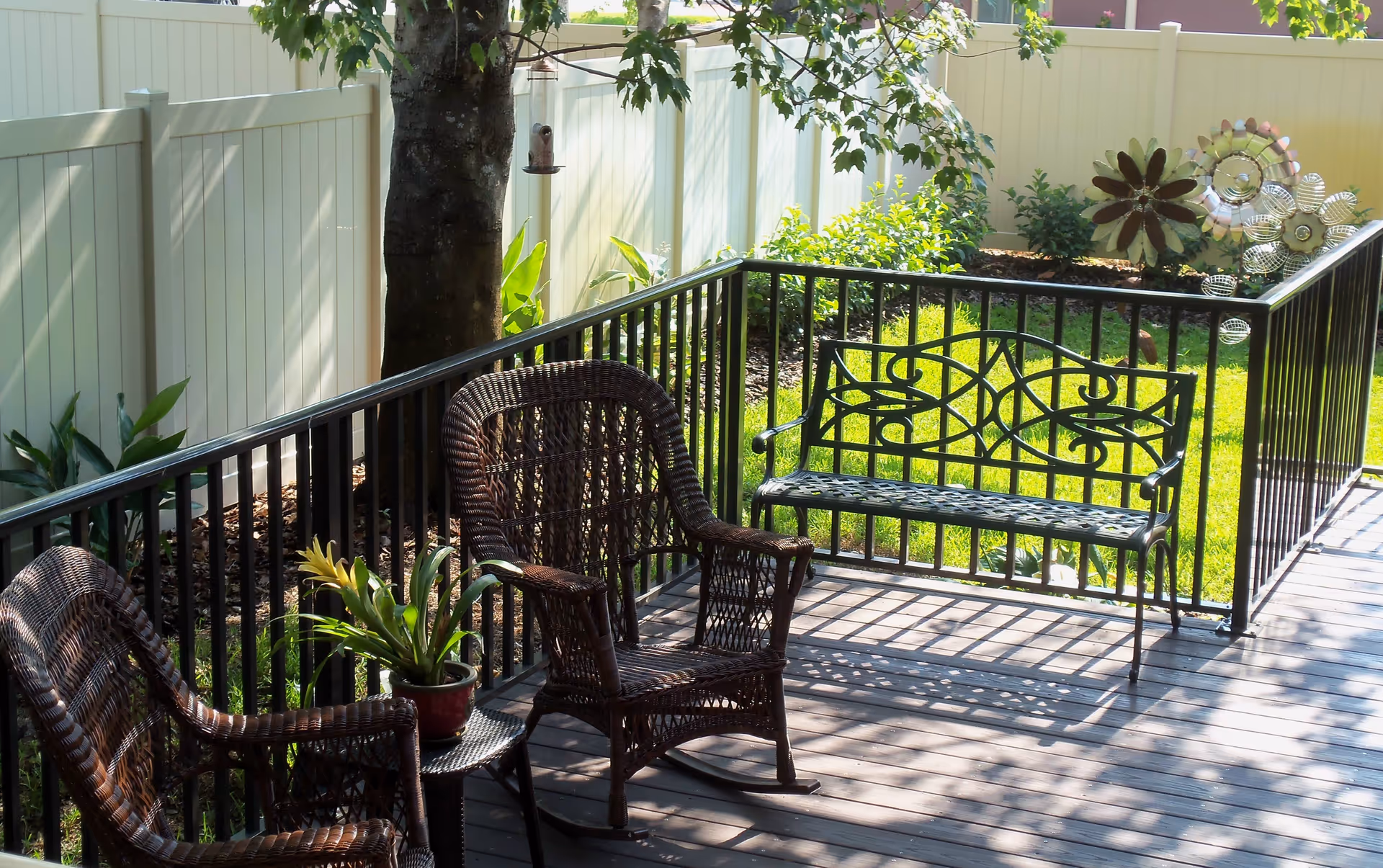 A sunlit outdoor patio area with two wicker chairs, a small table with a potted plant, and a decorative metal bench. The patio is surrounded by a black metal railing and a white privacy fence, with green grass and garden decorations in the background.