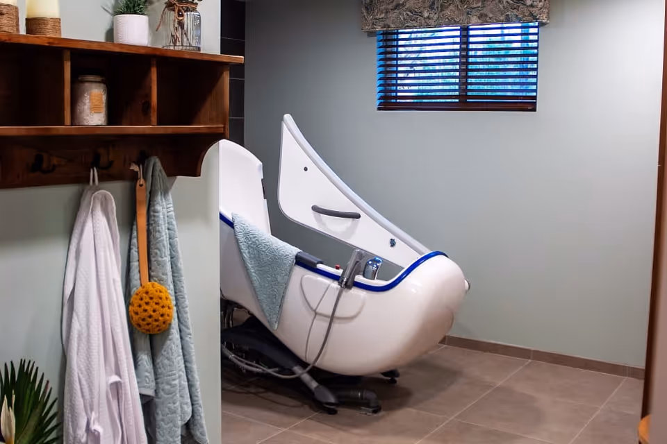 An accessible reclining bath chair in a tiled bathroom with towels hanging on a wooden wall shelf and a small window with blinds.