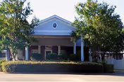 Front exterior view of Southern Springs Healthcare Facility showing a single-story building with a triangular pediment above the entrance, flanked by trees and bushes.
