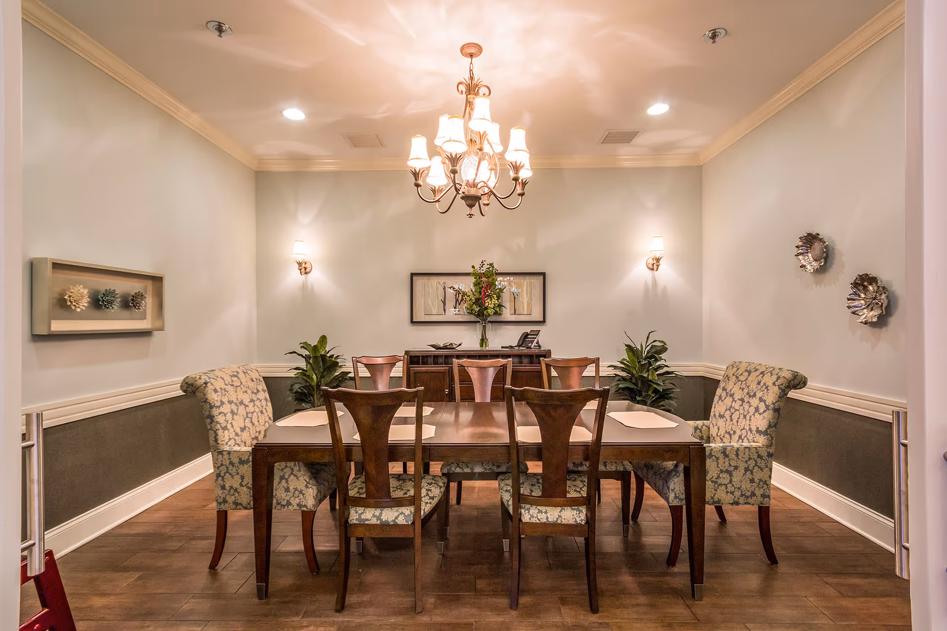 A formal dining room with a wooden dining table surrounded by six chairs, two of which have floral upholstery. A chandelier with multiple lights hangs from the ceiling, and two wall sconces provide additional lighting. There is a sideboard against the back wall with a vase of flowers and a framed artwork above it. Decorative wall art and plants are placed on the side walls.