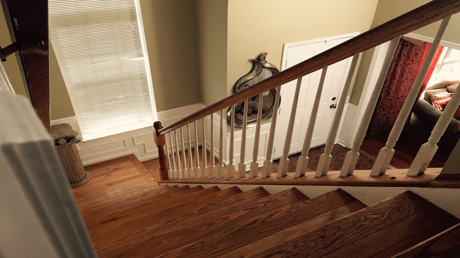 View looking down a wooden staircase with white balusters inside a home. At the bottom of the stairs is a white front door with a decorative mirror on the wall next to it. To the right, a living room area with a couch and red curtains is partially visible. A window with closed blinds is on the left side near the stairs.