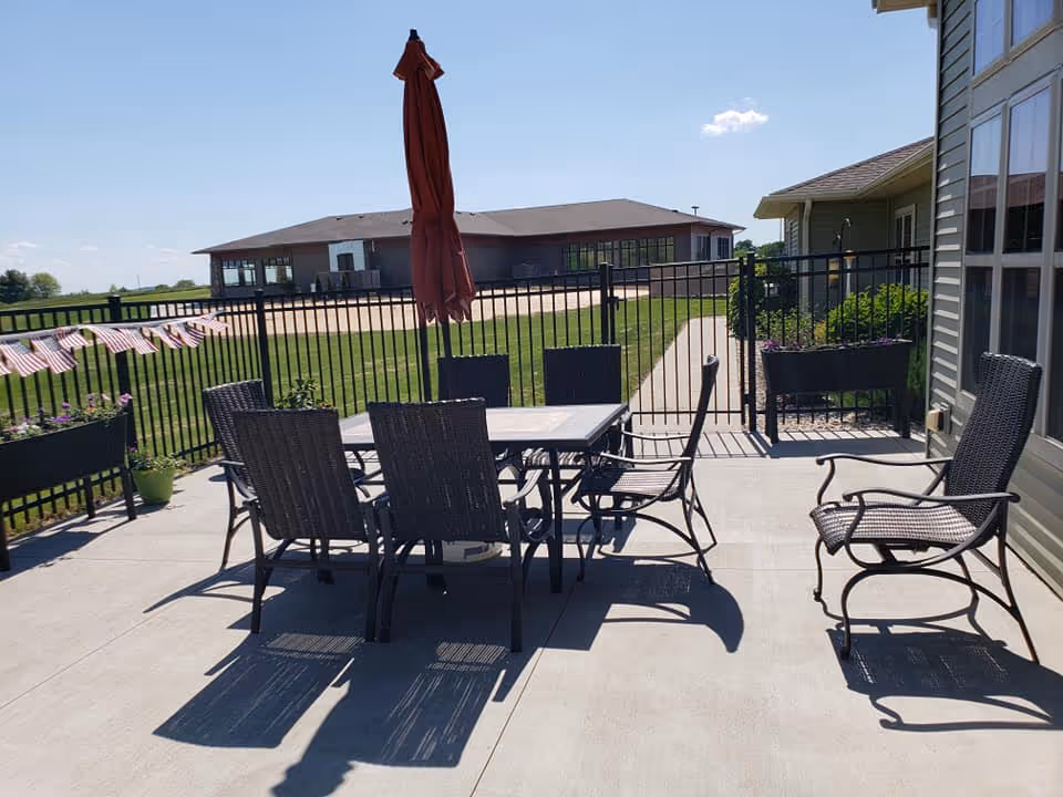 Outdoor patio area with a metal table and six matching chairs, one of which is slightly apart from the table. A closed red umbrella stands in the center of the table. The patio is surrounded by a black metal fence with a gate leading to a grassy area and a building in the background under a clear blue sky.