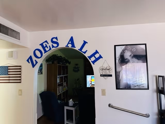 Interior view of a living space with a white wall featuring blue letters spelling 'ZOES ALH' above an arched doorway. To the left of the arch is a wooden American flag wall decoration. To the right of the arch is a framed black and white photo of a woman with hearts drawn above her head and a small decorative sign that says 'Welcome'. A grab bar is mounted on the wall below the photo. Inside the room beyond the arch, a bookshelf and a television are visible.