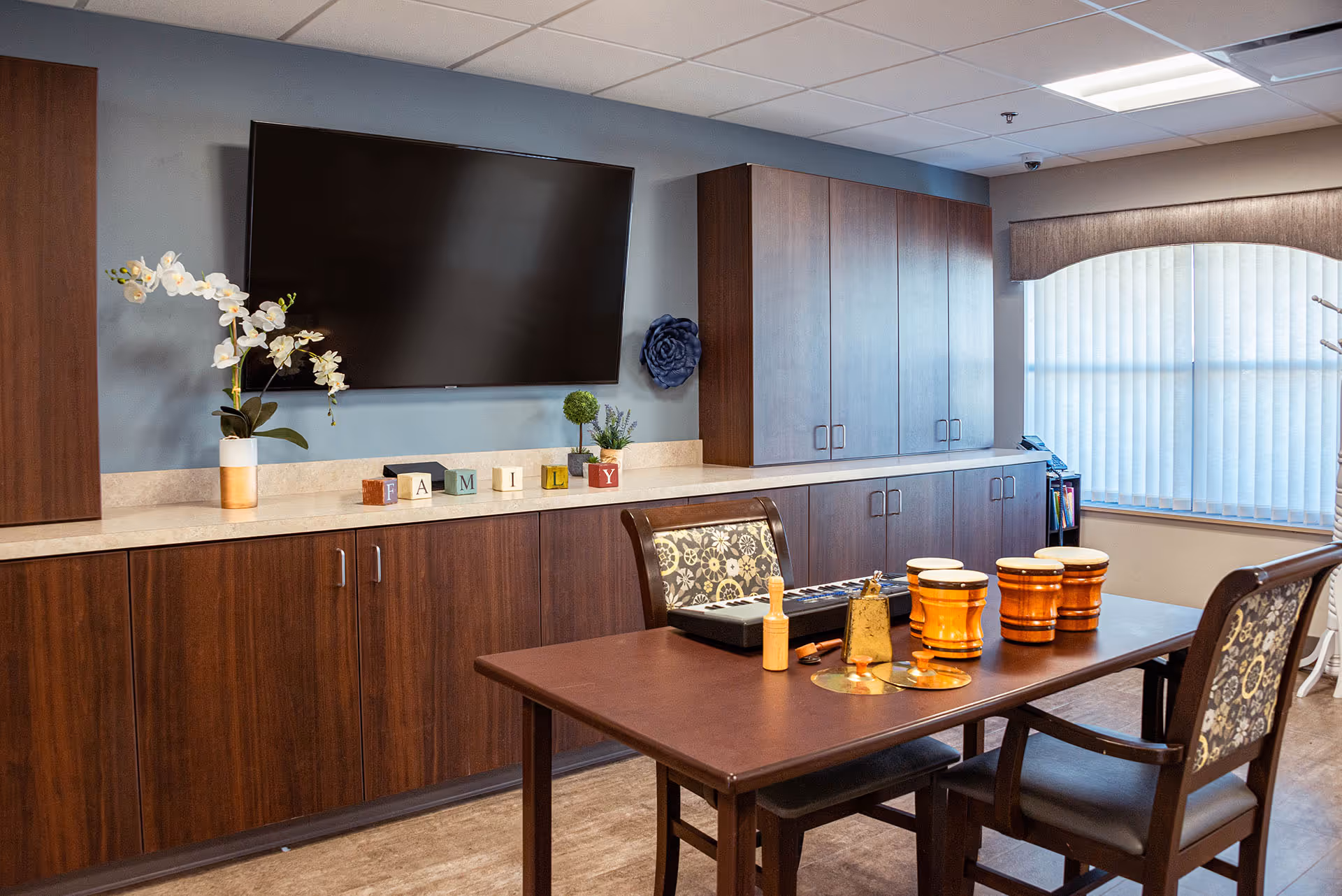 A cozy room with a wooden table and two chairs featuring floral upholstery. On the table are several percussion instruments including drums and a bell. Behind the table is a long countertop with dark wood cabinets above and below. A flat-screen TV is mounted on the wall above the counter, and decorative items including a white orchid plant and blocks spelling 'FAMILY' are displayed. A window with vertical blinds allows natural light into the room.