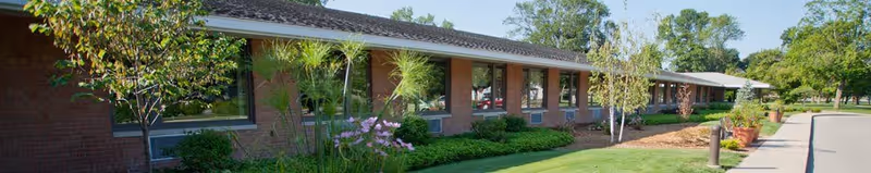 Exterior view of a single-story brick building with large windows, surrounded by well-maintained green shrubs, trees, and a grassy lawn under a clear sky.