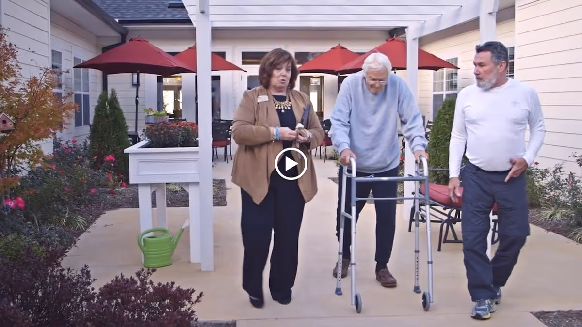 Three adults—one using a walker—walking through a covered outdoor courtyard with red umbrellas, planters, and seating.
