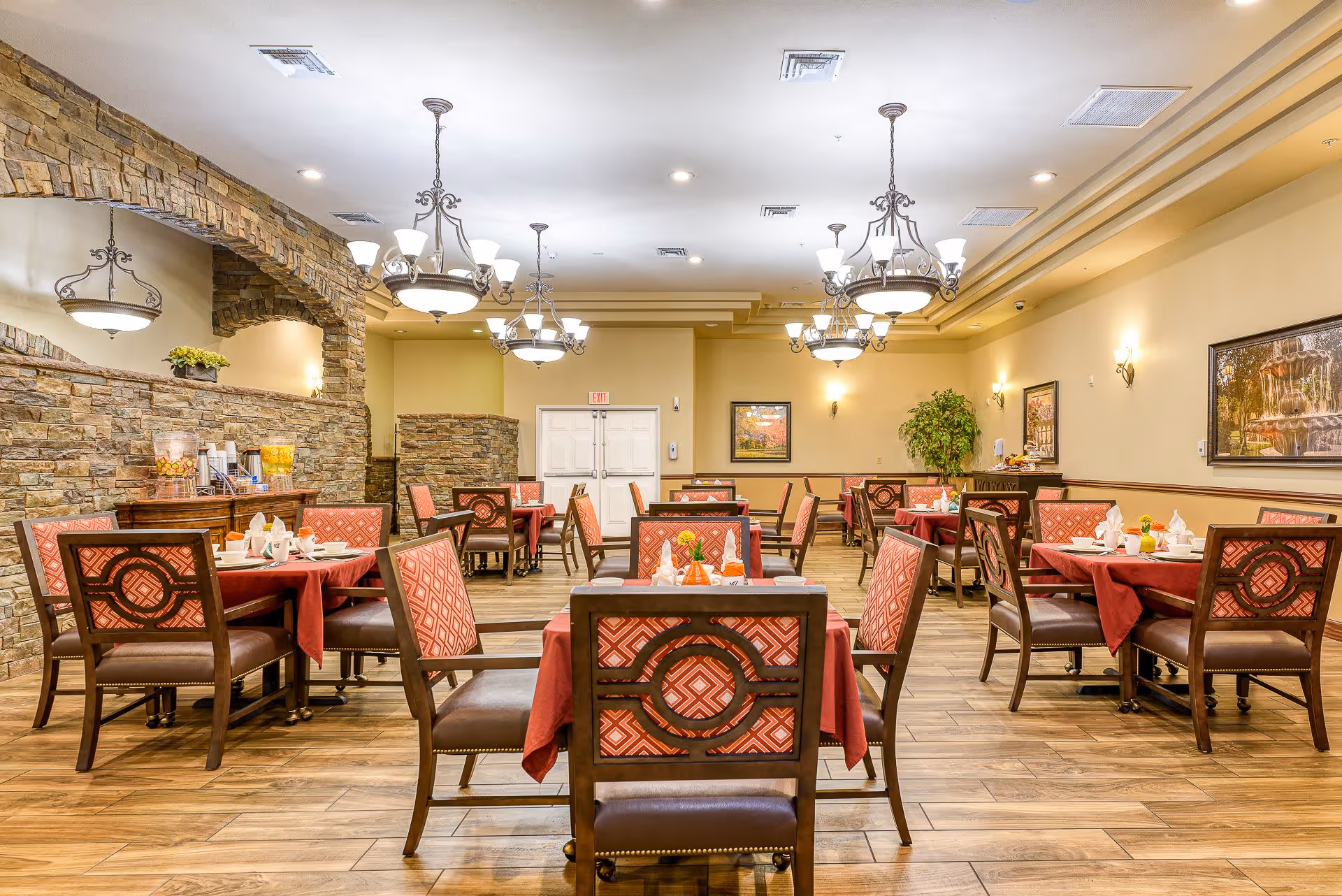A spacious dining room with multiple tables covered in red tablecloths and set with white napkins and dishes. The chairs have wooden frames with red patterned upholstery. The room features warm lighting from chandeliers and wall sconces, stone accent walls, wooden flooring, and framed artwork on the walls.