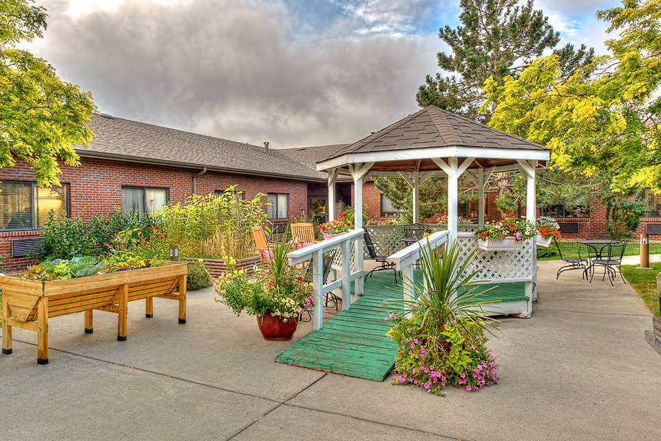 Outdoor courtyard area at Life Care Center of Casper featuring a white gazebo with a green ramp, surrounded by potted plants and flowers. There are wooden raised garden beds with various plants, patio tables and chairs, and a brick building in the background under a cloudy sky.