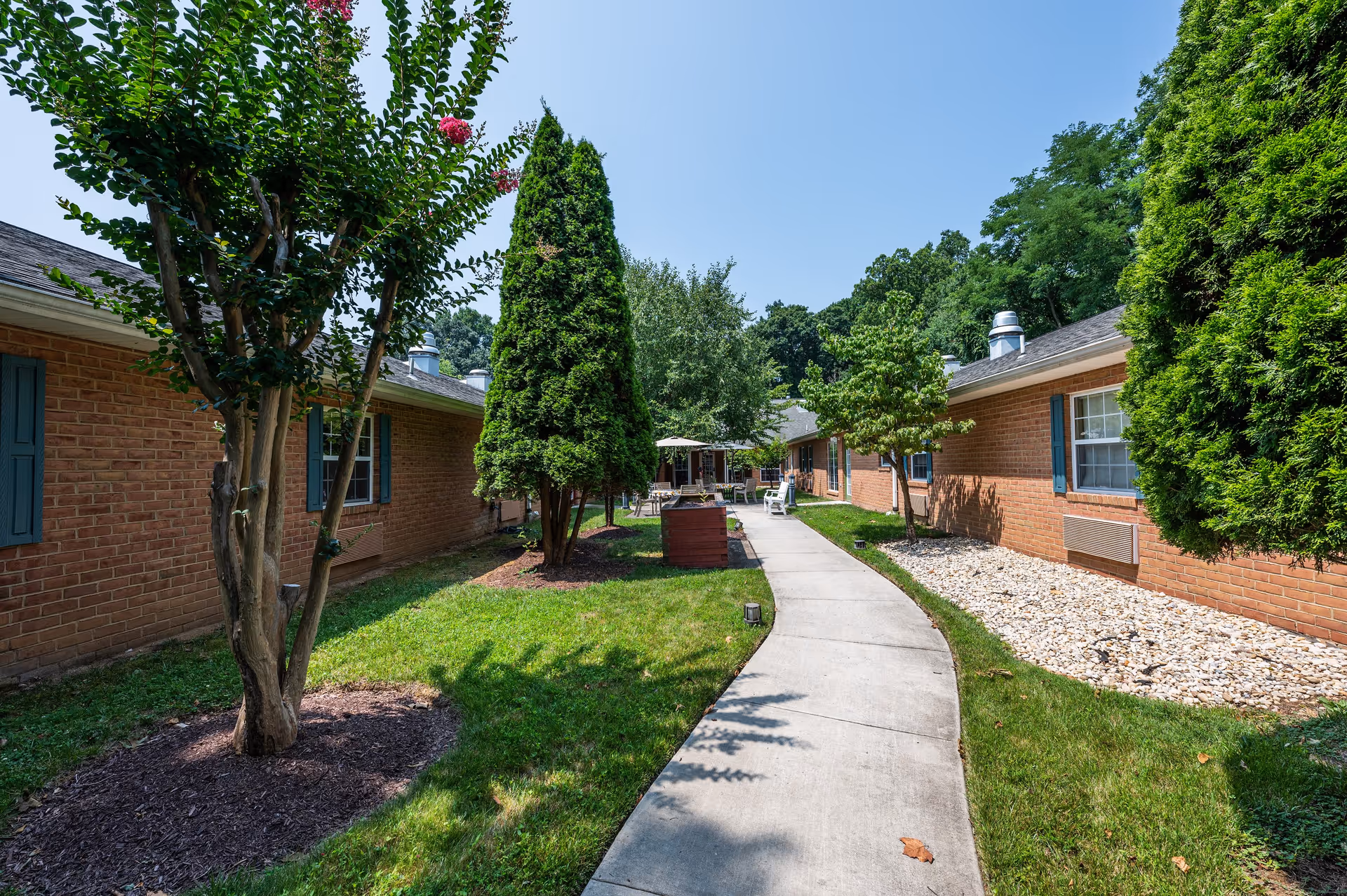 A sunny outdoor pathway between two single-story brick buildings with blue window shutters. The pathway is surrounded by green grass, trees, and shrubs, leading to a seating area with tables and chairs in the distance under umbrellas.