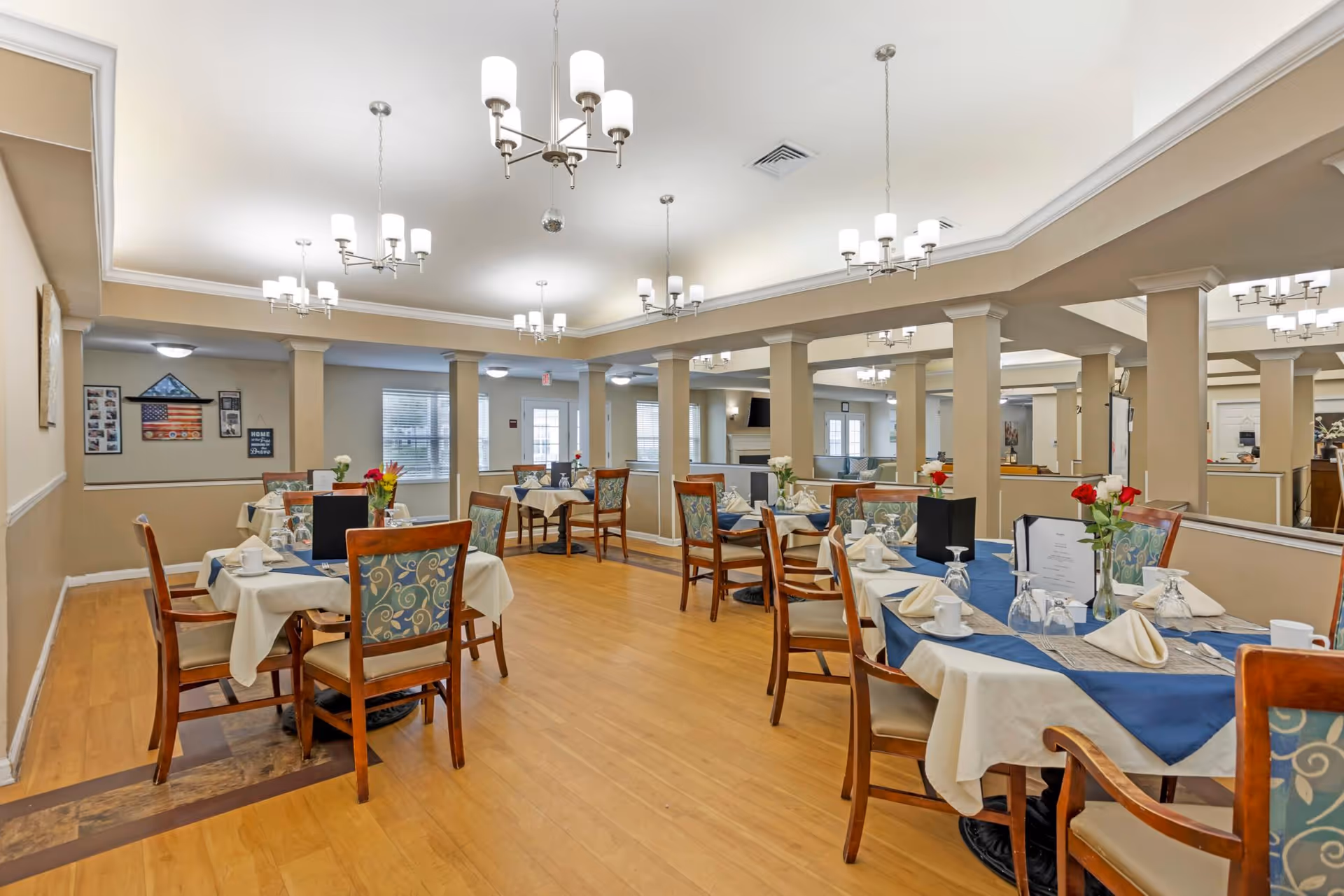 Spacious dining room with multiple set tables, wooden chairs, chandeliers, and place settings.