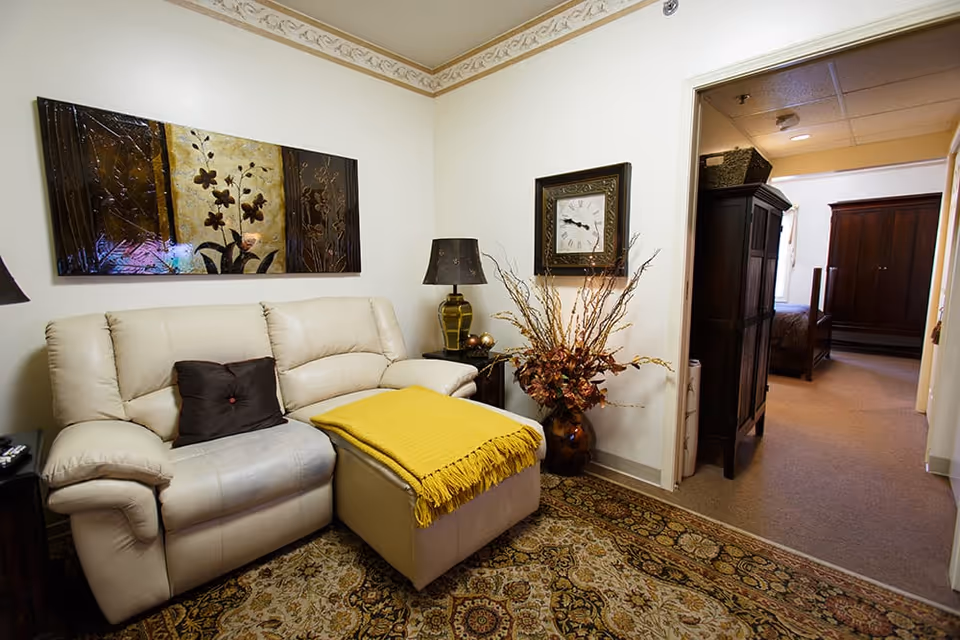 A cozy living room area with a cream-colored leather loveseat, a brown pillow, and a matching ottoman covered with a yellow throw blanket. There is a decorative floral painting on the wall above the loveseat, a table lamp on a side table, a large vase with dried branches and flowers, and a wall clock. The room has a patterned carpet and an open doorway leading to a bedroom with dark wooden furniture.