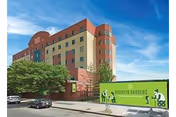 Exterior view of a multi-story building with a red and beige facade under a blue sky. In front of the building, there is a green sign that reads 'Brooklyn Gardens Nursing and Rehabilitation Center' with illustrations of people and trees. A few cars are parked along the street beside the building.