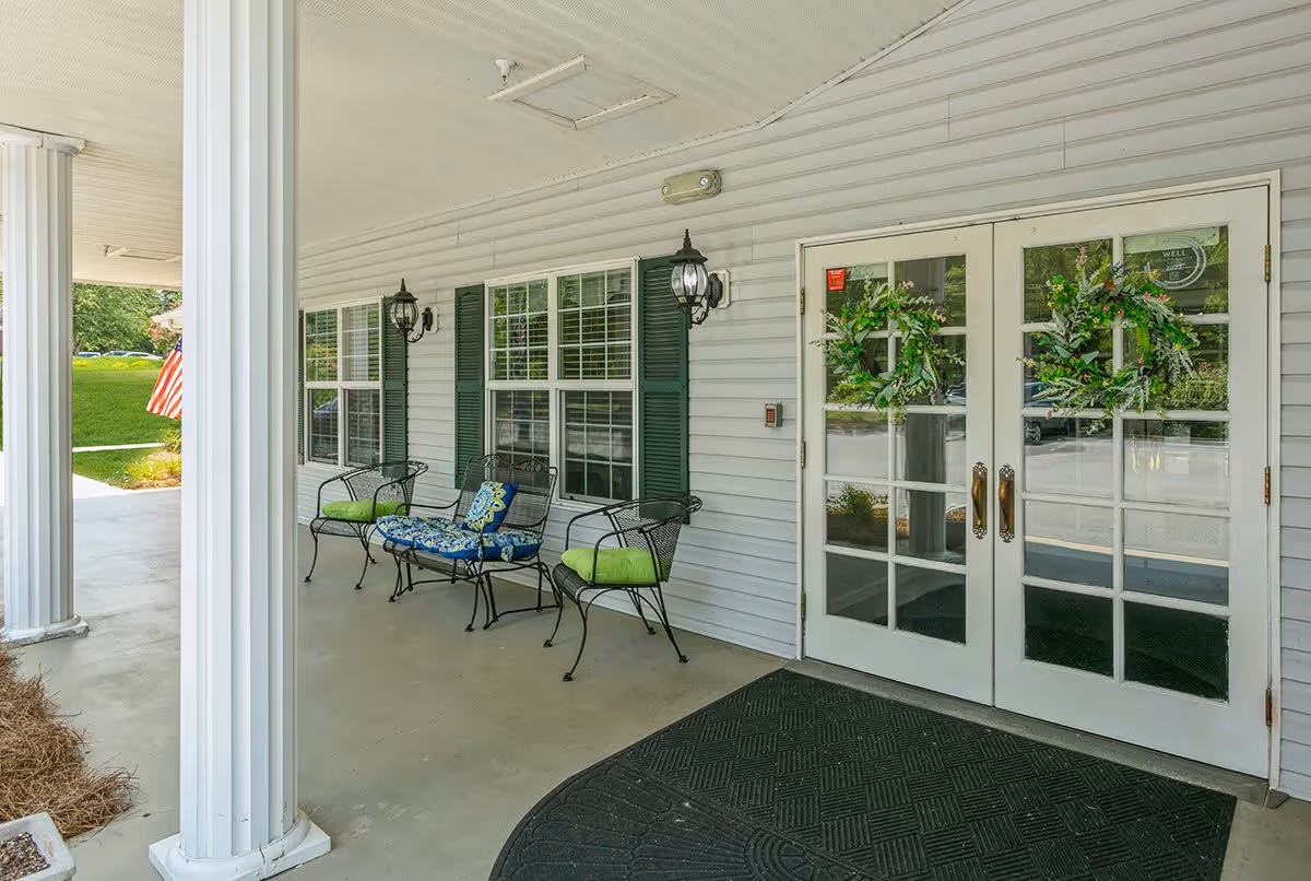 Covered porch area with white columns, two black metal chairs with green cushions, a black metal bench with blue and green cushions, two wall-mounted lantern-style lights, and double glass doors decorated with green wreaths. An American flag is visible in the background on the left side.