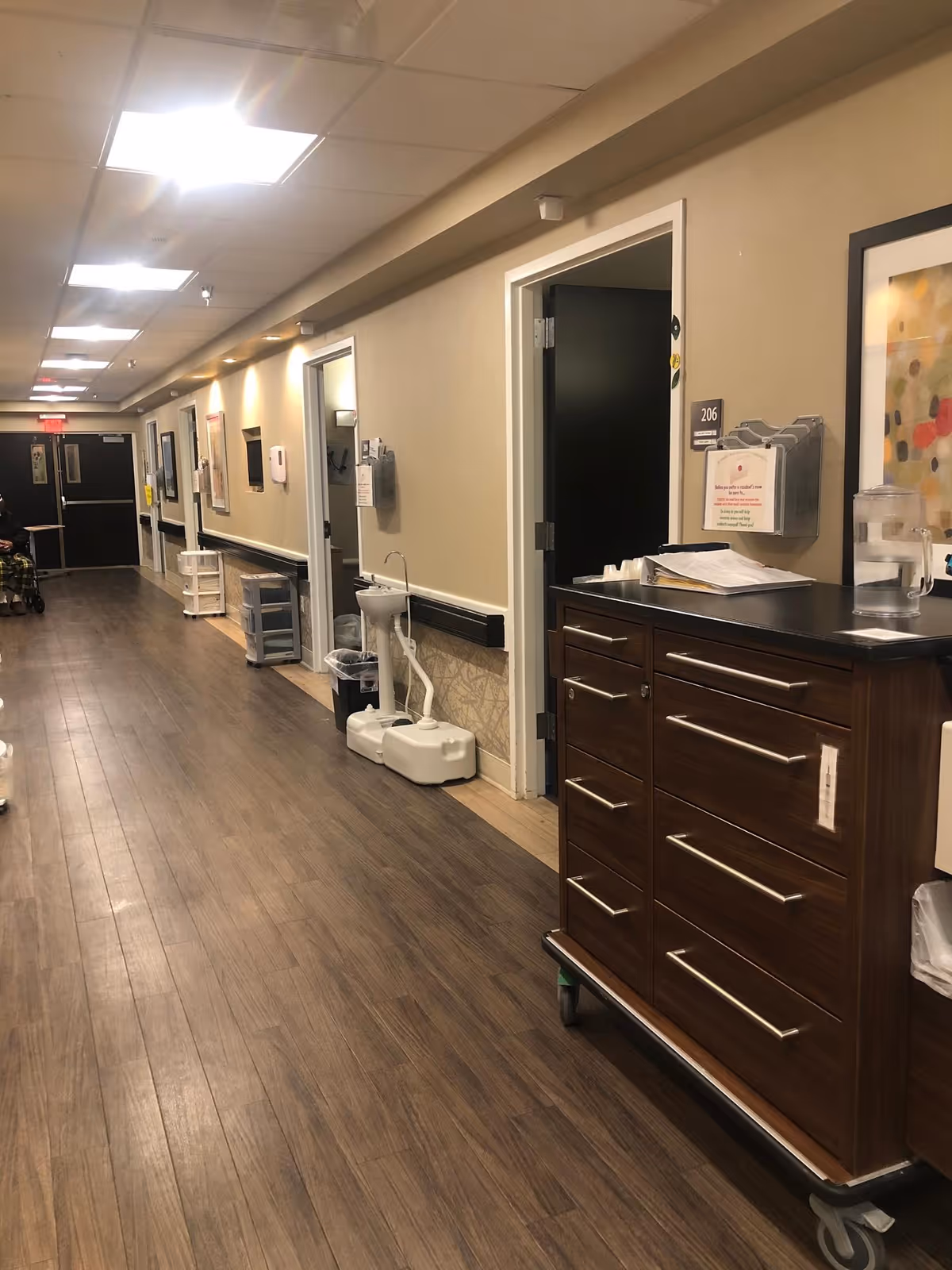 Long interior hallway of a senior living facility with wood floors, open room doors, and a mobile medical supply cart.