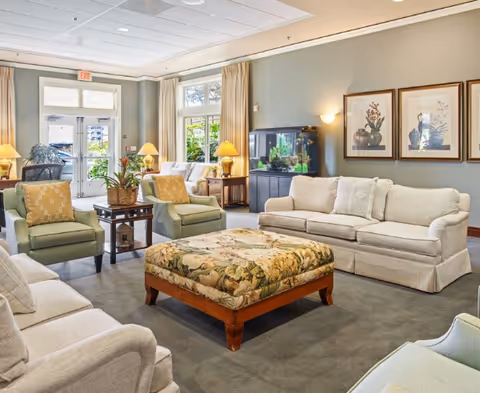 Bright furnished communal living room with sofas, armchairs, a patterned ottoman, lamps, framed art, and an aquarium.