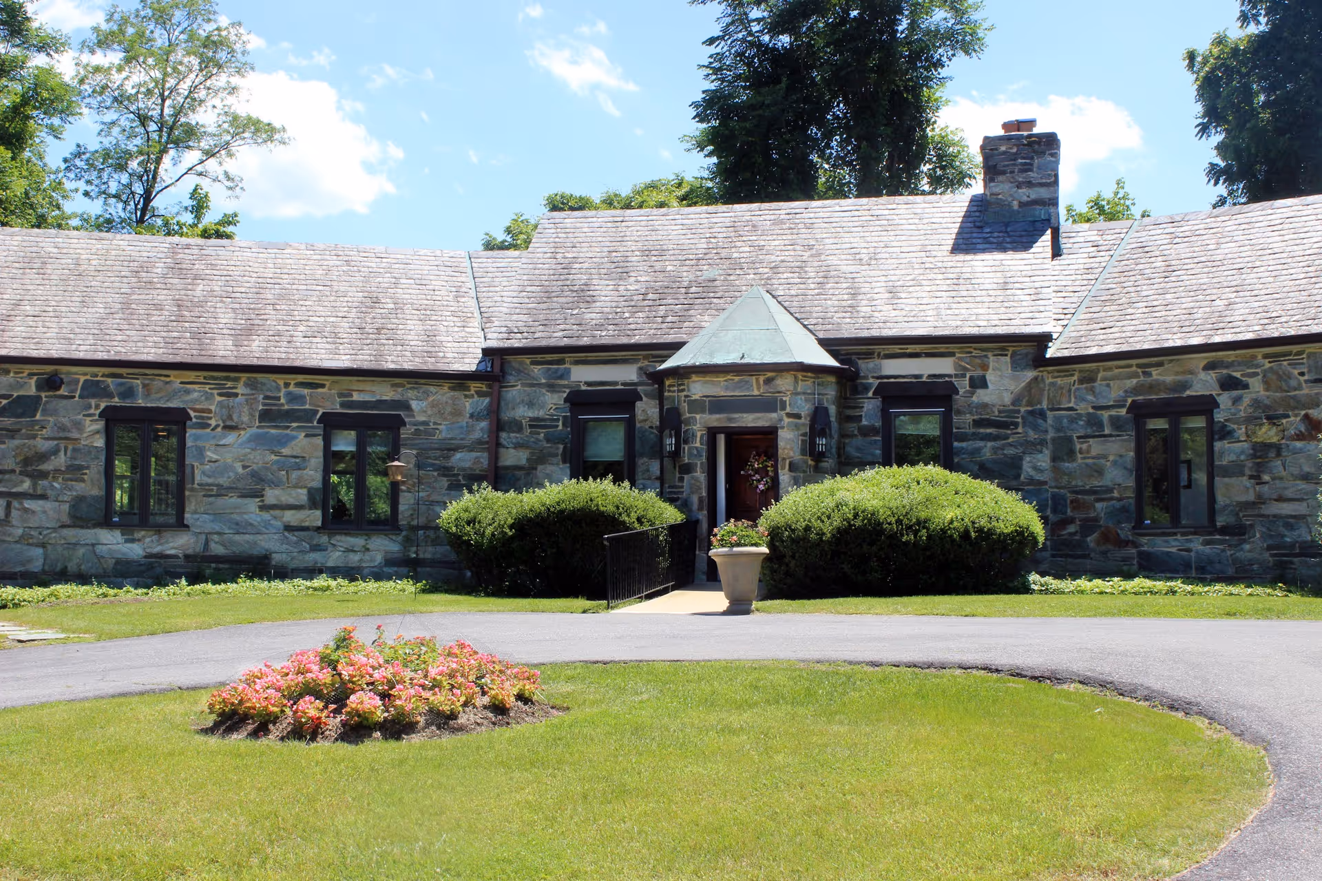 Stone building with a shingled roof and several windows, surrounded by green bushes and a circular driveway with a flower bed in the center, under a blue sky with some clouds.