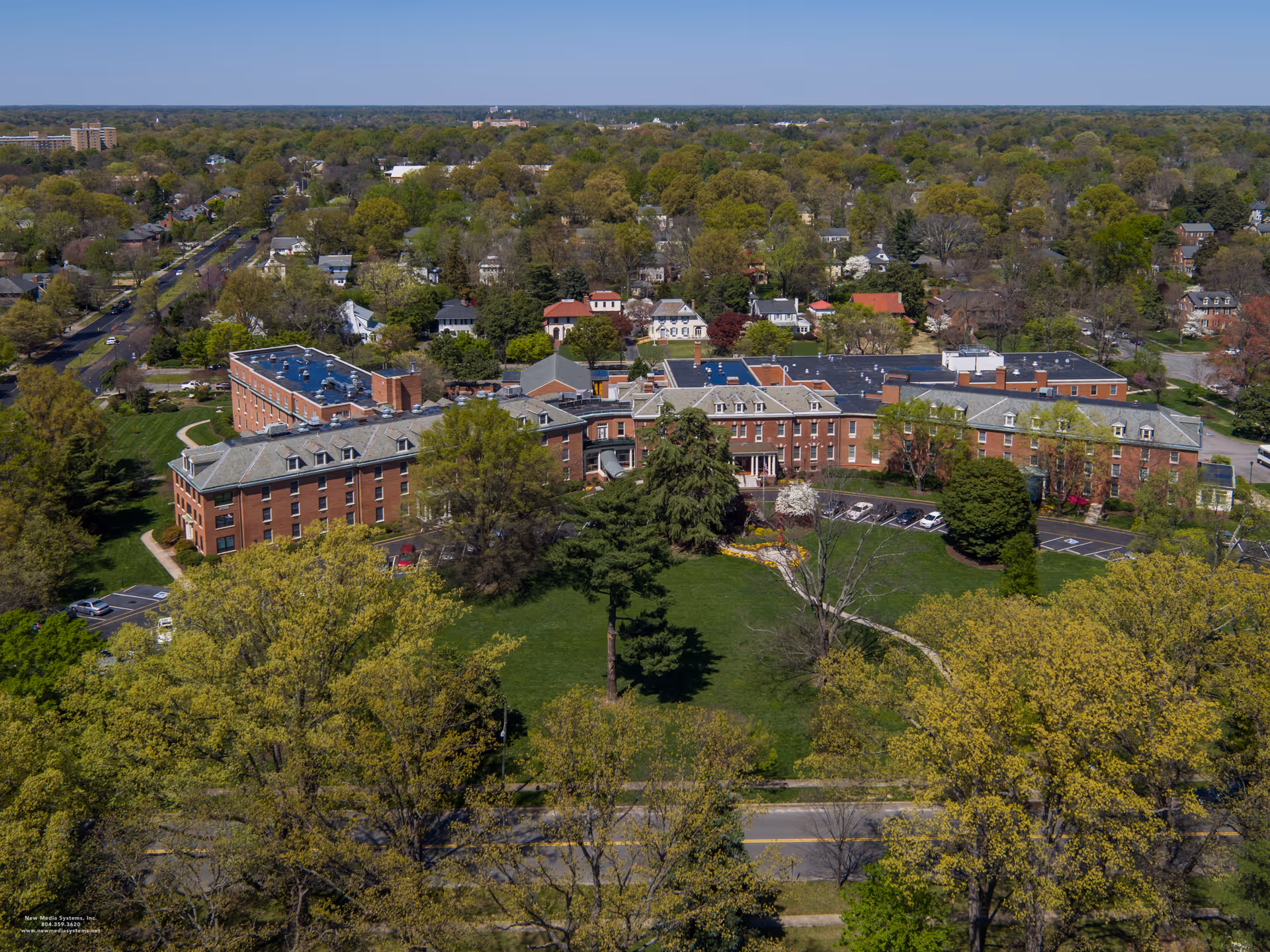 Aerial view of Hermitage Richmond, a large brick senior living facility surrounded by green lawns, trees, and a residential neighborhood with houses and streets in the background under a clear blue sky.