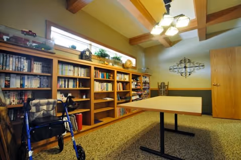A cozy room with wooden bookshelves filled with books and decorative items along one wall. A blue walker is positioned near the shelves. A rectangular table with a light wood top and black legs is centered in the room. The ceiling features wooden beams and a ceiling light fixture with multiple bulbs. The room has carpeted flooring and a closed wooden door on the right side.