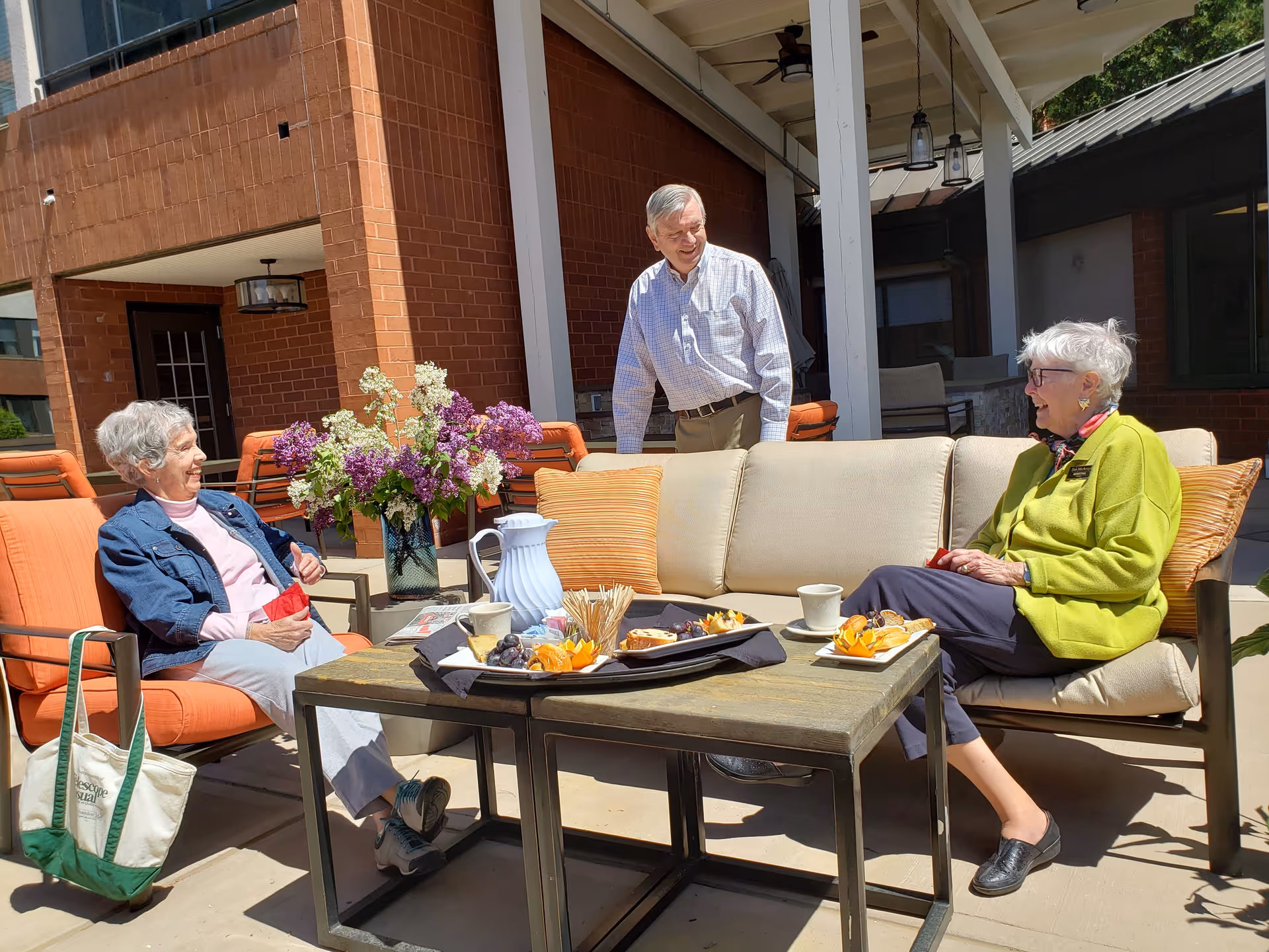 Three elderly people enjoying a sunny day on an outdoor patio. Two women are seated on cushioned chairs around a table with plates of snacks and cups, while a man stands behind the sofa smiling. The setting includes a brick building, a vase with flowers on the table, and comfortable outdoor furniture.