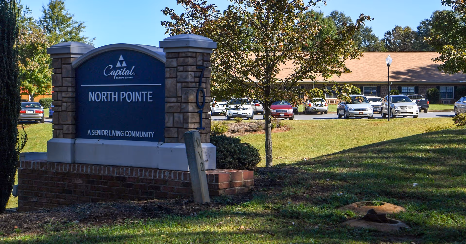 Outdoor view of the entrance sign for North Pointe, a senior living community by Capital Senior Living, with a parking lot and single-story building in the background surrounded by trees and grass.