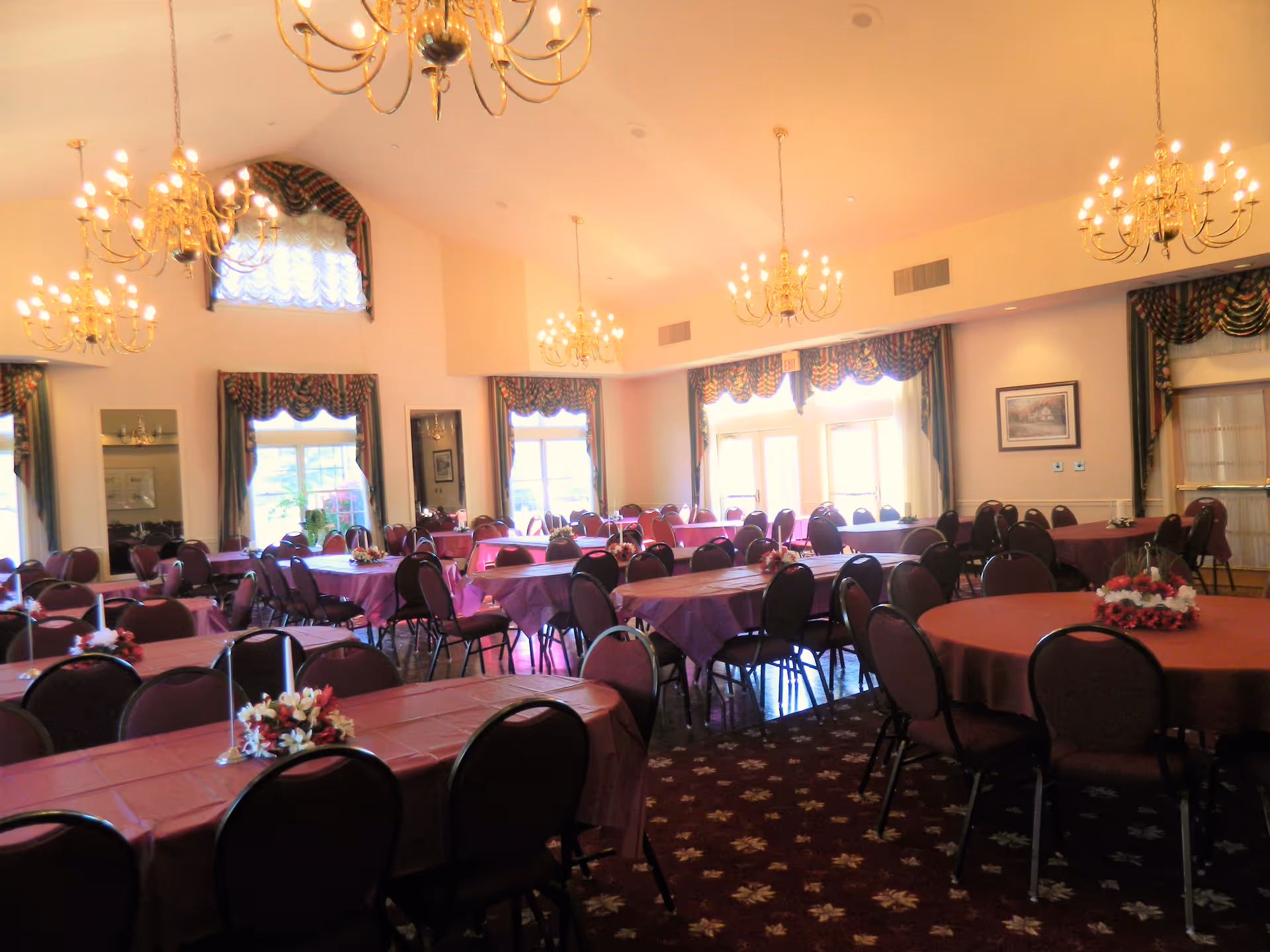 A spacious dining room with multiple round and rectangular tables covered with maroon tablecloths. Each table is surrounded by chairs and decorated with floral centerpieces and candles. The room features large windows with patterned curtains, high ceilings with several ornate chandeliers, and a carpeted floor with a floral pattern.
