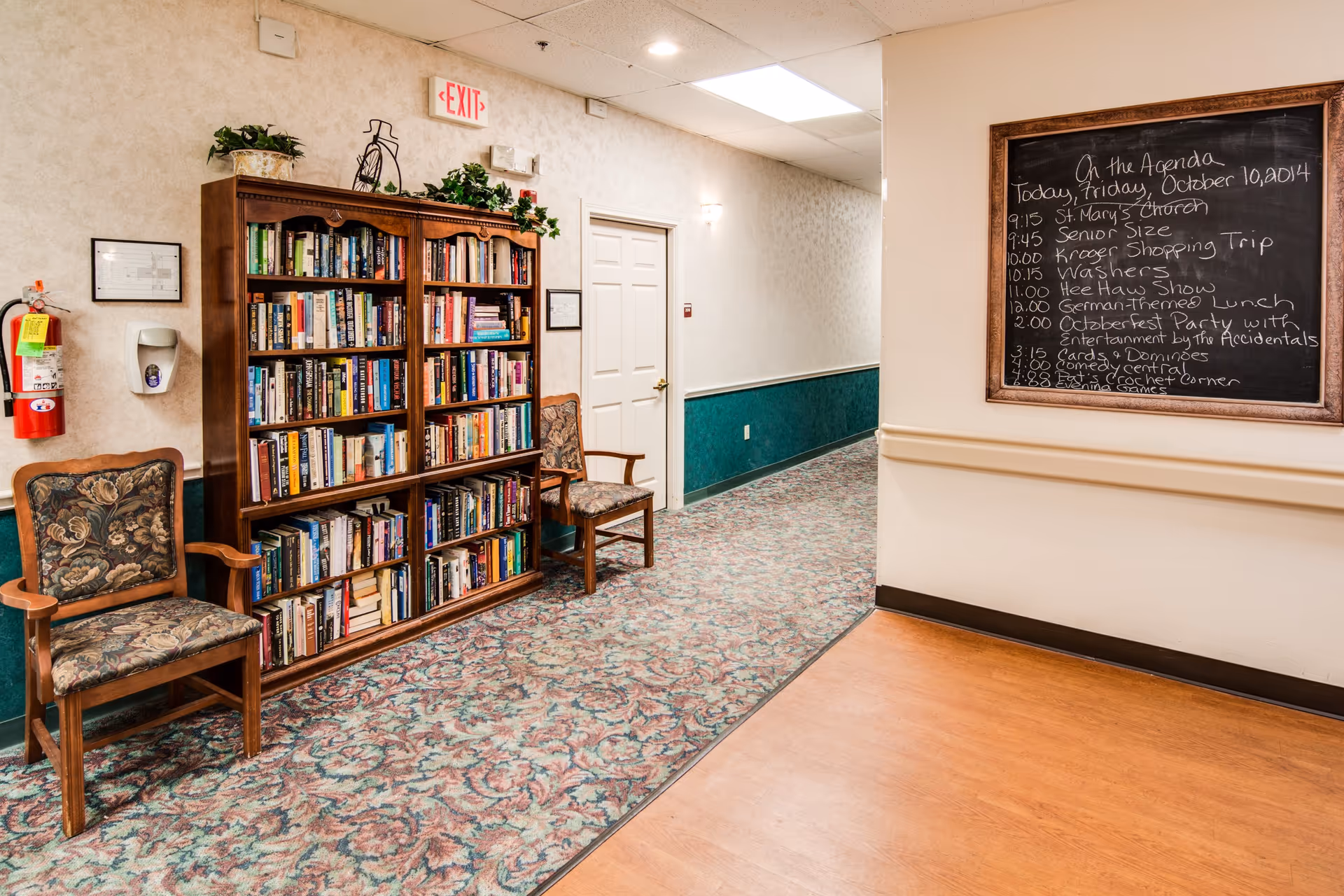 Interior hallway of a senior living facility with a wooden bookshelf filled with books, two upholstered chairs on either side of the bookshelf, a fire extinguisher mounted on the wall, and a chalkboard displaying the day's agenda. The hallway has patterned carpet and beige walls with a green wainscot.